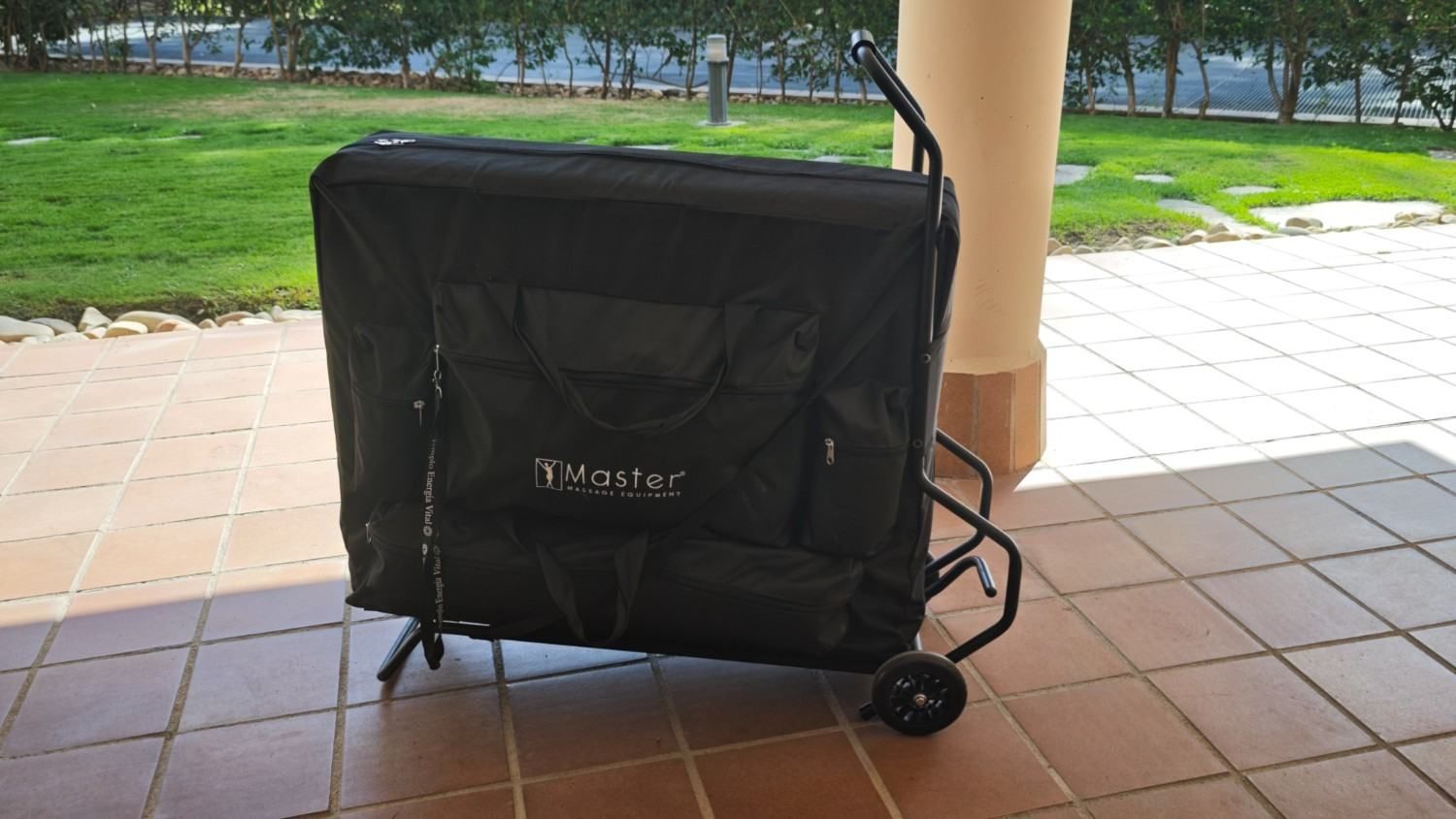 Carrito plegable negro con ruedas o cochecito cubierto en un patio embaldosado junto a una columna, con césped al fondo