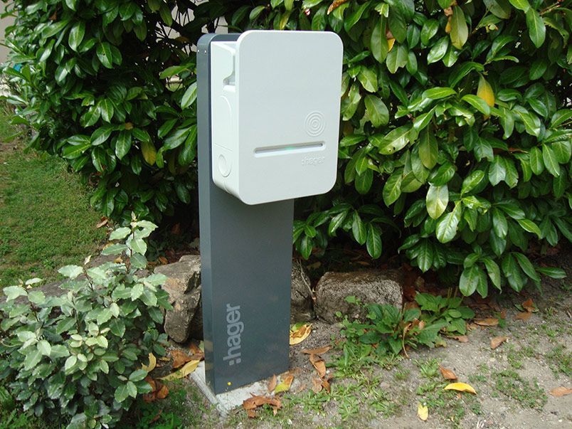 Borne de recharge grise pour véhicules électriques dans un jardin agrémenté de végétation verte. Le chargeur se compose d'un boîtier blanc et d'un support gris foncé.