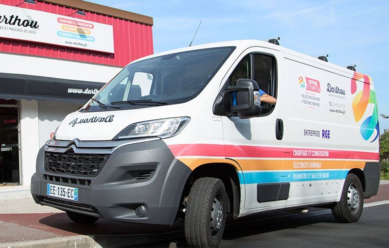 Une camionnette blanche arborant un logo coloré est garée devant un bâtiment. Le conducteur est visible.