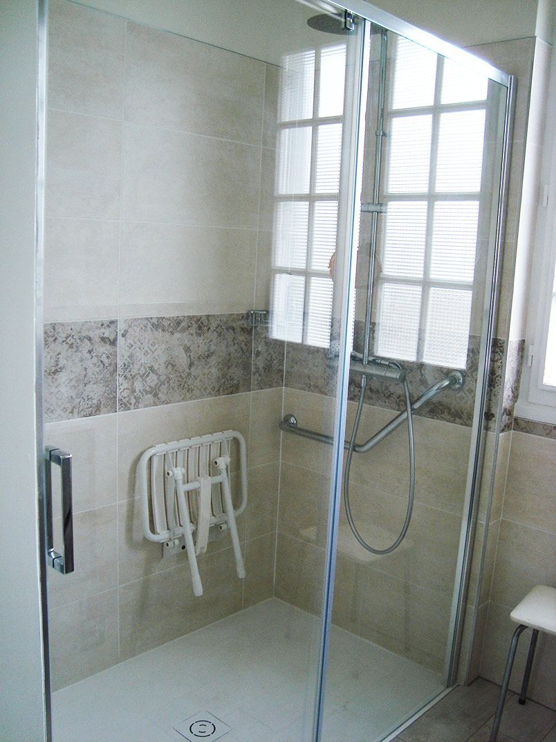 Douche accessible avec siège rabattable, barre d'appui et porte en verre, murs carrelés et fenêtre.