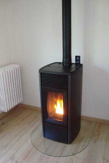 Poêle à granulés noirs avec flammes, cheminée noire et protection de sol en verre sur un plancher en bois.