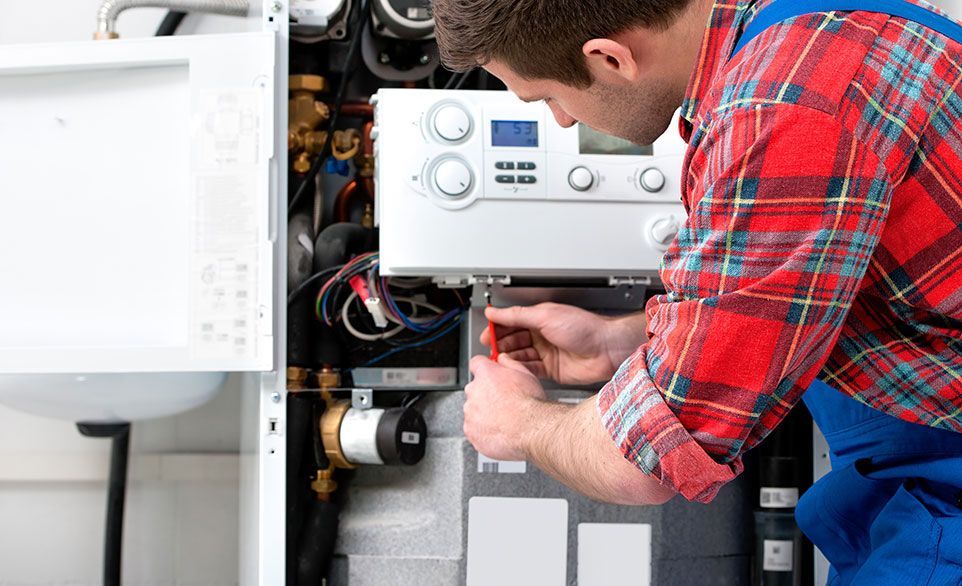 Une personne vêtue d'une salopette bleue et d'une chemise à carreaux rouges répare une chaudière à l'aide d'un tournevis.
