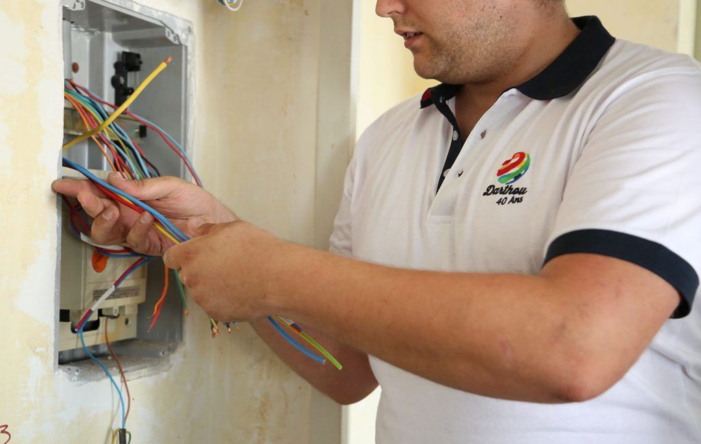 Un homme en chemise blanche travaille sur le câblage électrique d'une boîte murale, tenant des fils de couleurs différentes.