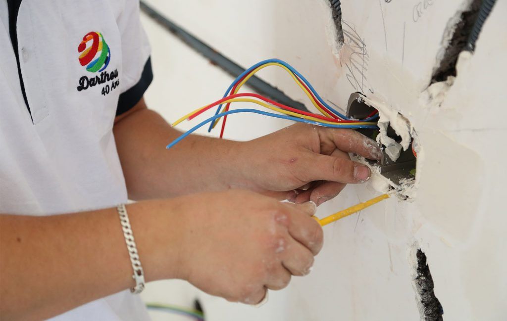 Une personne installe des câbles électriques dans une prise murale. Tournevis jaune, fils bleus, rouges et jaunes.