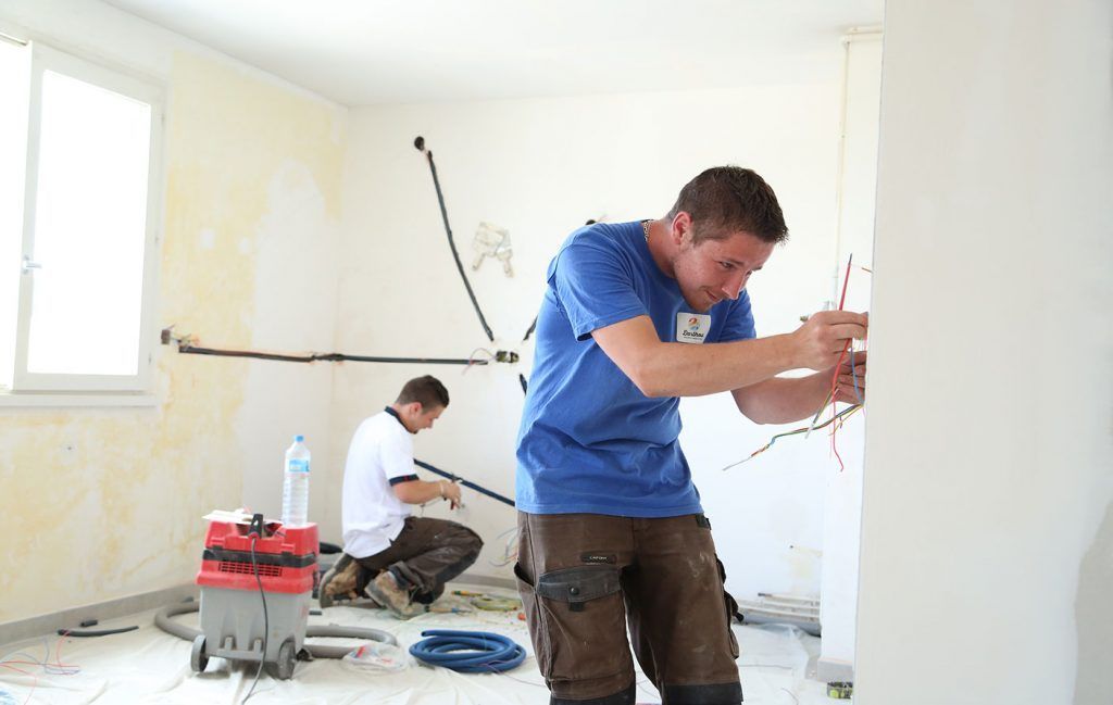 Deux ouvriers installent des câbles électriques dans une pièce aux murs apparents.