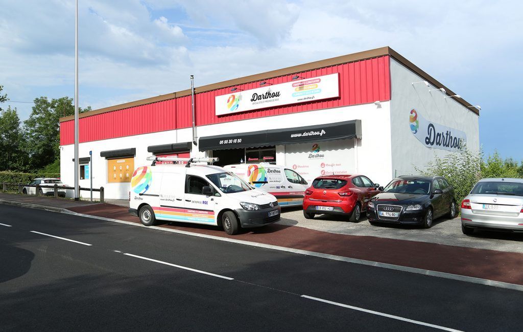 Façade commerciale rouge et blanche, véhicules stationnés et camionnette de livraison.