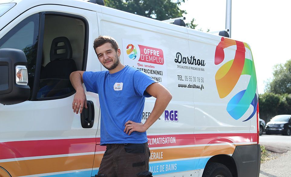 Un homme en chemise bleue s'appuie sur une camionnette blanche ornée d'un logo arc-en-ciel, dans un cadre extérieur ensoleillé.