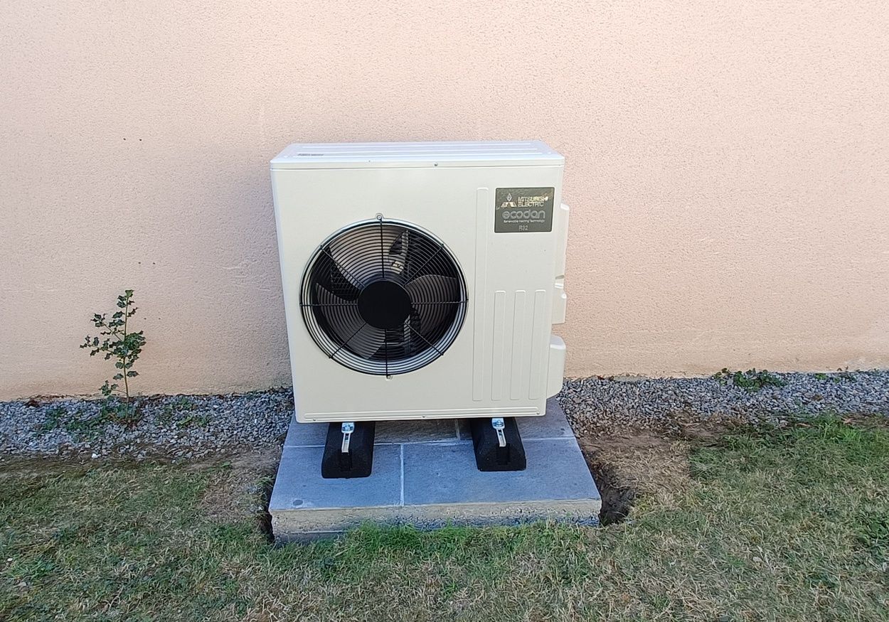 Unité de climatisation extérieure contre un mur beige, sur une plateforme grise dans une cour gazonnée.