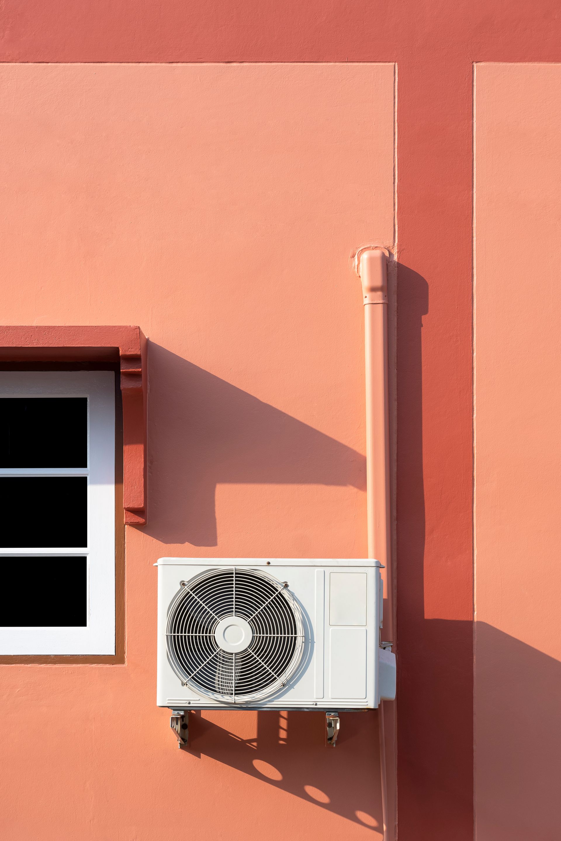 Climatiseur blanc fixé sur la façade d'un bâtiment couleur corail, près d'une fenêtre et d'une gouttière.