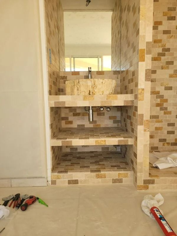 Coin lavabo de la salle de bain avec carrelage en pierre. Miroir, étagères ouvertes et tuyauterie apparentes.