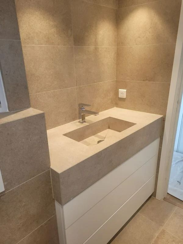 Salle de bains avec lavabo rectangulaire, plan de travail gris et meuble-lavabo blanc. Murs carrelés beiges. Robinet chromé.
