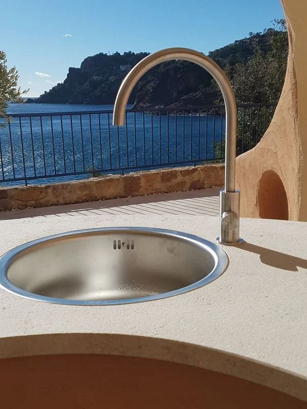 Évier en acier inoxydable avec robinet arqué, encastré dans un plan de travail en pierre, avec vue sur la mer et les montagnes.