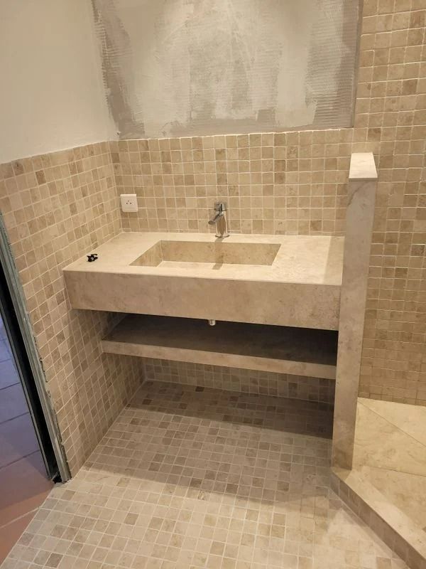 Salle de bains avec carrelage beige, lavabo en pierre avec robinet et deux étagères.