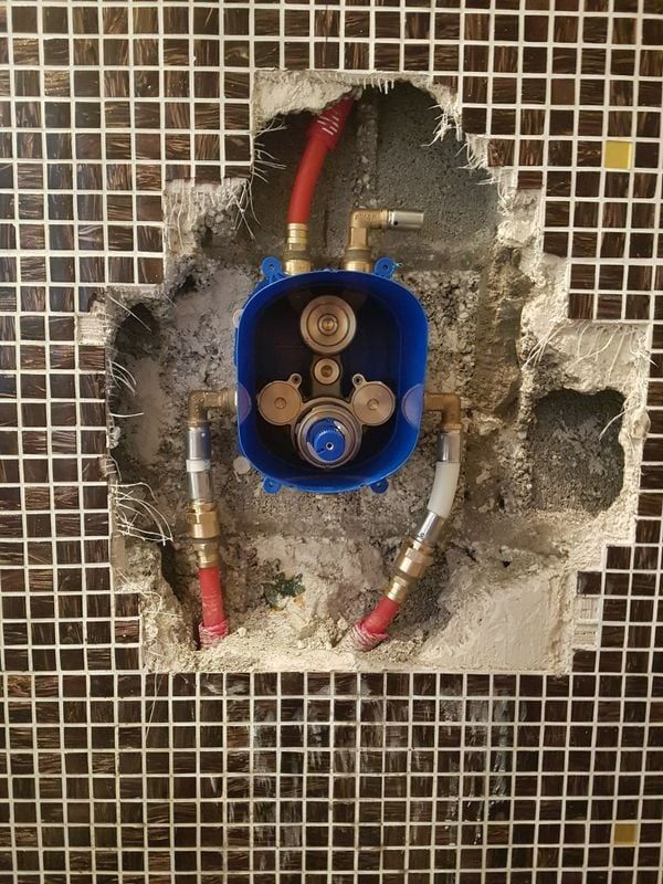 Boîtier de robinetterie de douche bleu installé dans un mur carrelé ; plomberie apparente et tuyaux rouges/blancs.