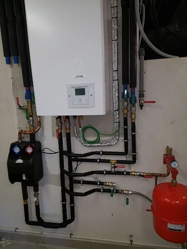 Une chaudière murale blanche avec tuyaux et manomètres, un système de pompe noir et un vase d'expansion rouge sont installés sur un mur blanc.