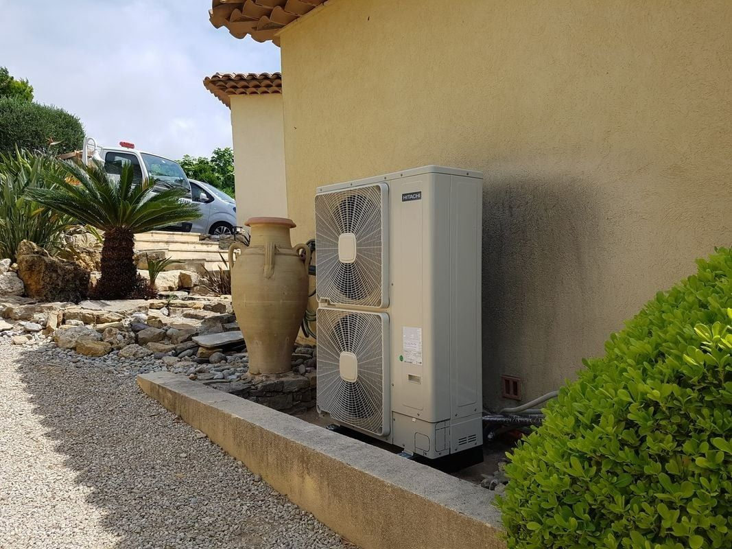 Unité de pompe à chaleur à côté d'un bâtiment, gravier et plantes à l'extérieur.