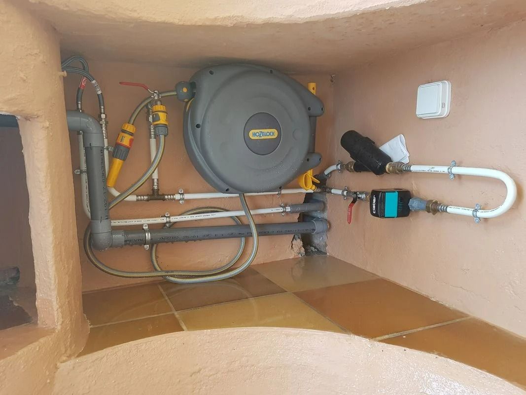 Enrouleur de tuyau d'arrosage et plomberie dans une alcôve extérieure. Les couleurs dominantes sont le beige et le gris.