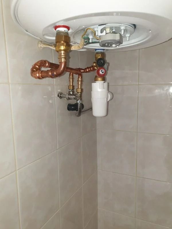 Tuyaux et vannes en cuivre raccordés à un chauffe-eau blanc dans une salle de bains carrelée.