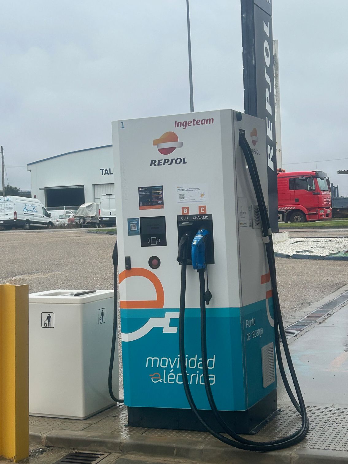 Gasolinera con columnas, surtidores y panel de control en blanco y negro. Señalización naranja y blanca.