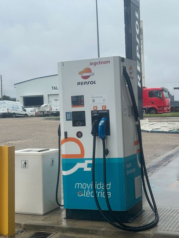 Gasolinera con columnas, surtidores y panel de control en blanco y negro. Señalización naranja y blanca.