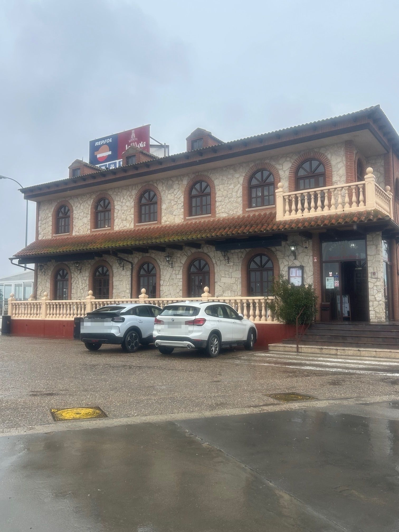Edificio de ladrillo de dos pisos con ventanas arqueadas, un balcón y un cartel en el techo; dos autos estacionados en el frente en un día lluvioso.