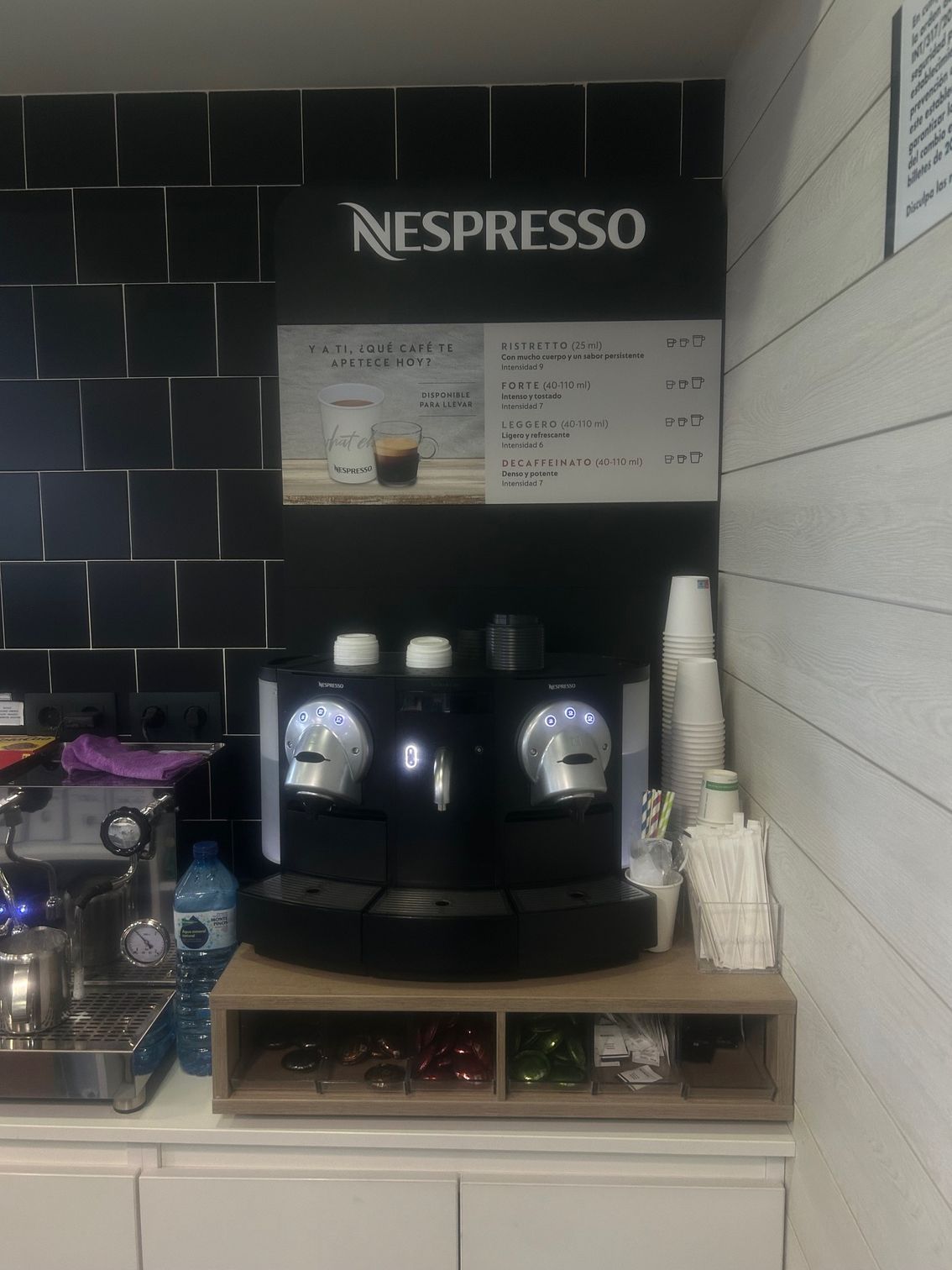 Cafetera Nespresso con menú, junto a máquina de café espresso, tazas y agitadores en un mostrador.