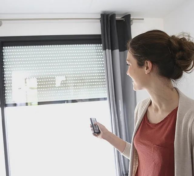 Una mujer sostiene un control remoto frente a una ventana.