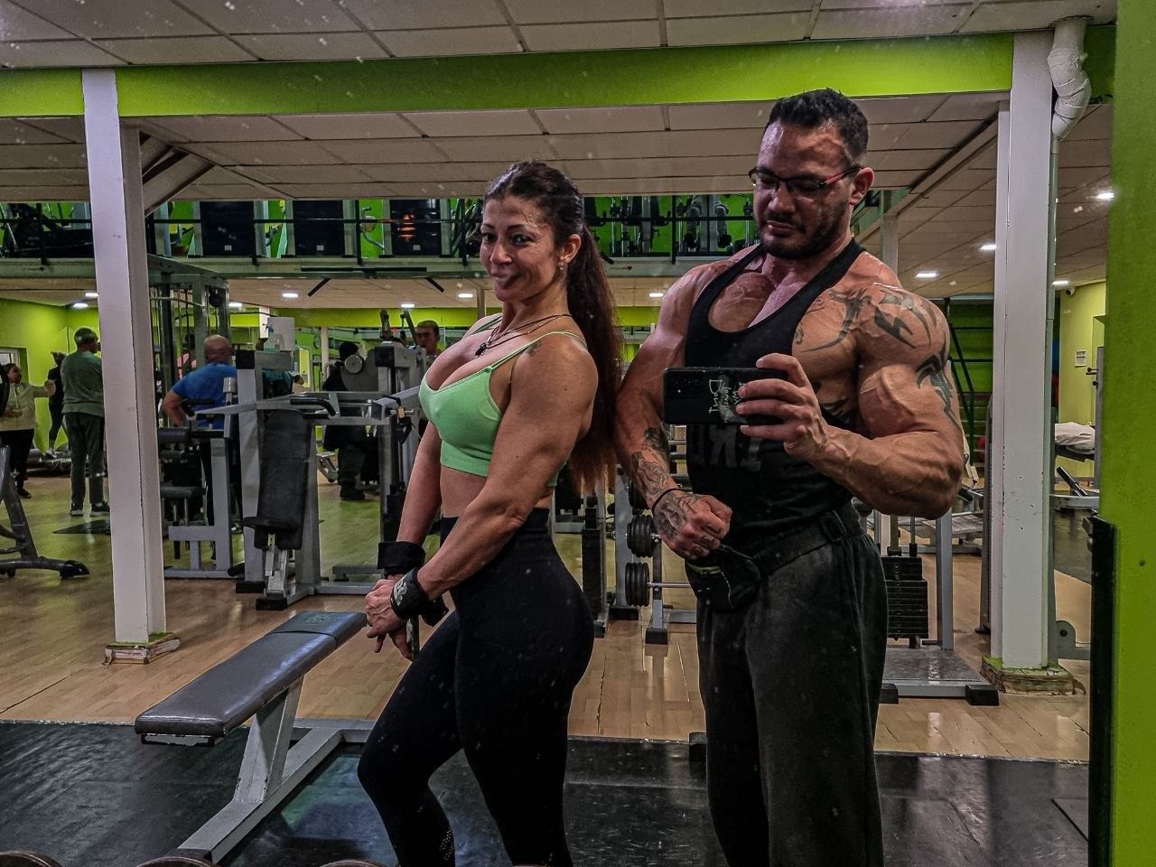 Dos personas que van al gimnasio hacen ejercicio flexionando sus músculos frente a los espejos, vestidas con ropa deportiva