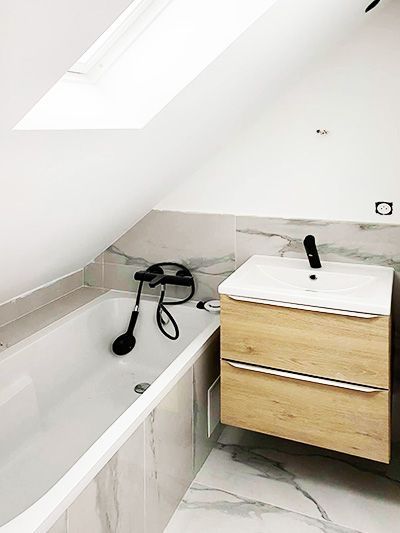 Salle de bains avec baignoire blanche, meuble-lavabo en bois clair et fenêtre dans le plafond mansardé.