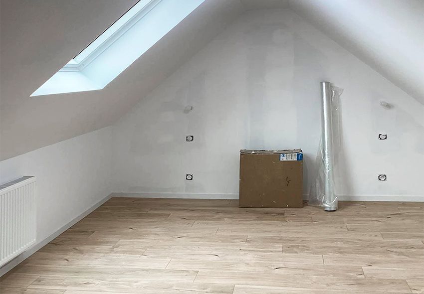 Chambre mansardée avec parquet, murs blancs et puits de lumière. Un carton et des objets emballés s'y trouvent.