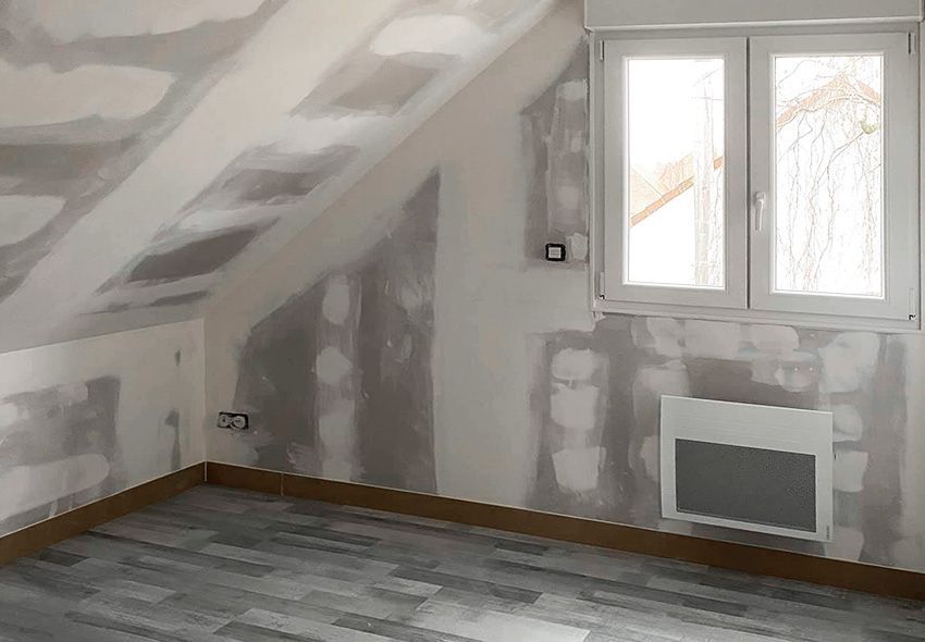 Pièce en travaux avec plafond en pente, murs réparés, fenêtre et sol gris.