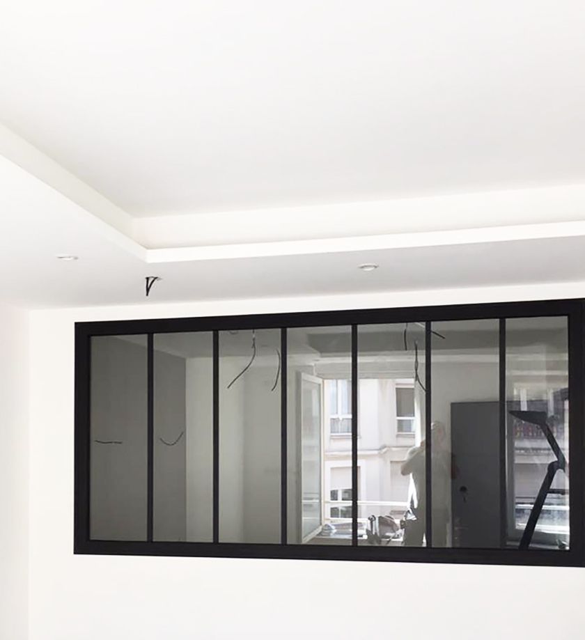 Fenêtre à cadre noir sur un mur blanc. Les vitres reflètent un intérieur et un immeuble avec des balcons.