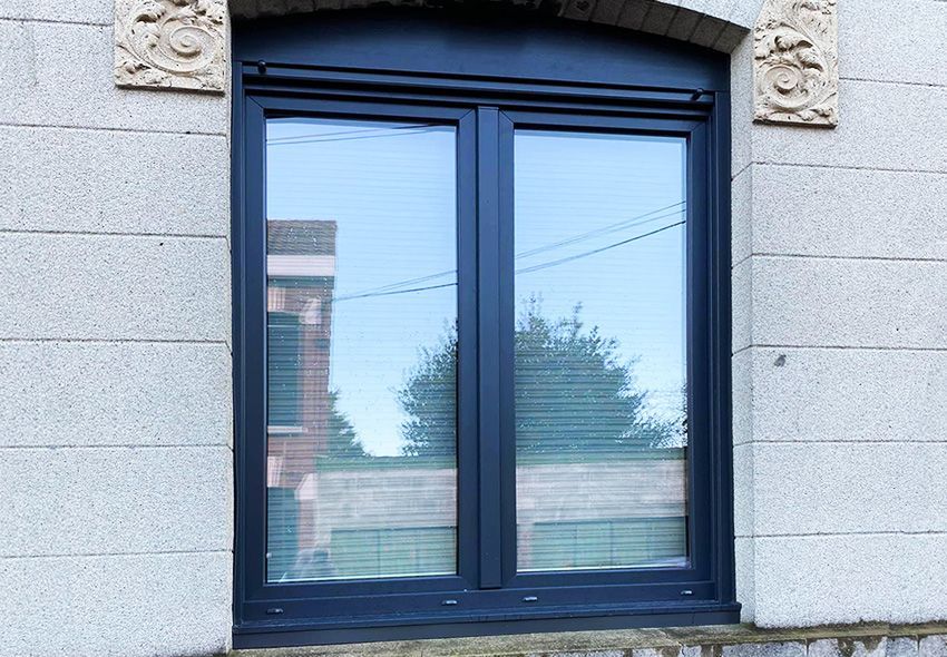 Fenêtre à cadre noir avec stores, reflétant les bâtiments et le ciel, encastrée dans une façade en pierre.