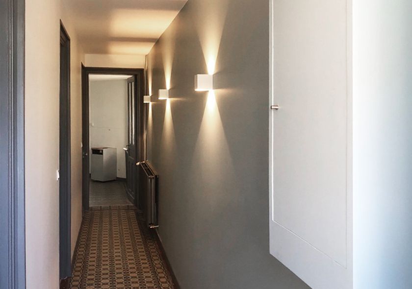 Couloir étroit aux murs gris, appliques murales cubiques, armoire blanche et sol carrelé à motifs.