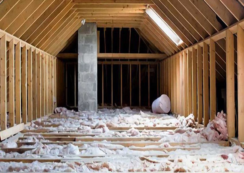 Grenier non aménagé avec charpente en bois, isolation, cheminée et éclairage fluorescent.