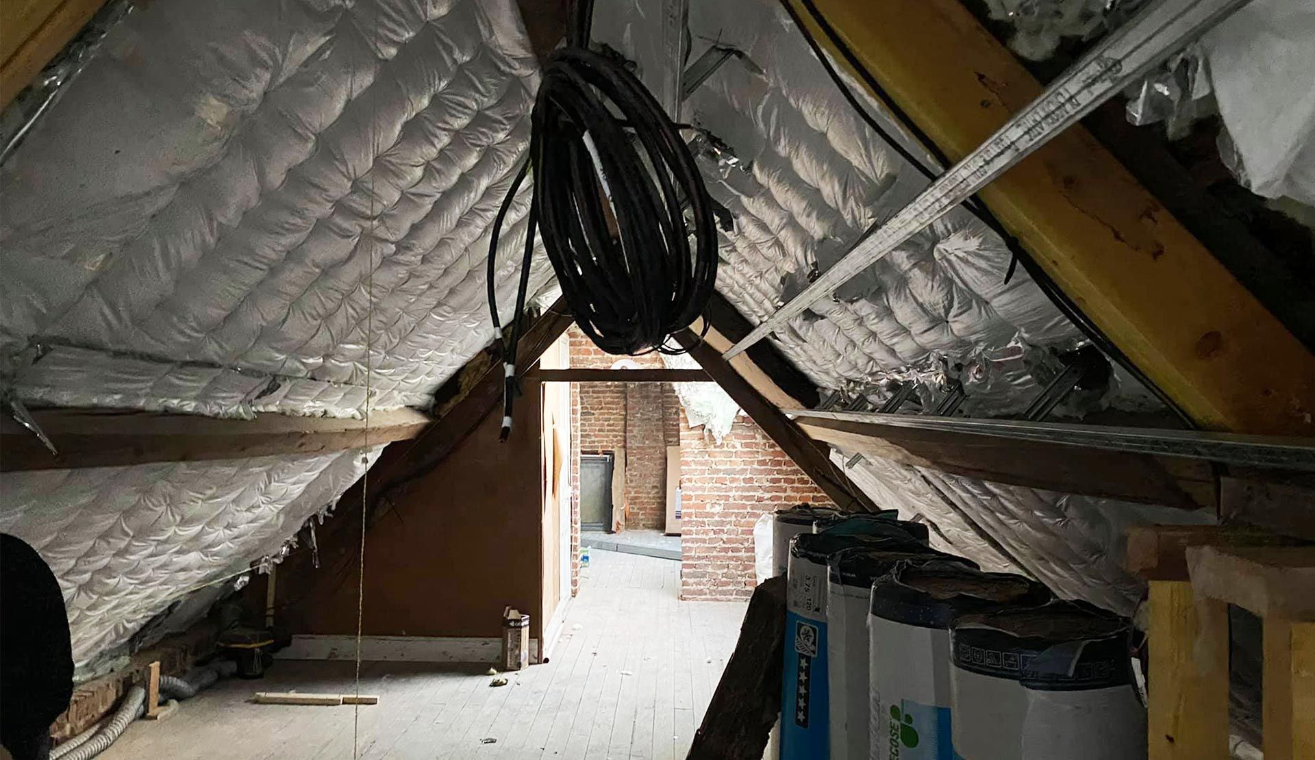 Intérieur du grenier avec isolation, câblage, poutres en bois et une vue sur l'extérieur à travers une porte.