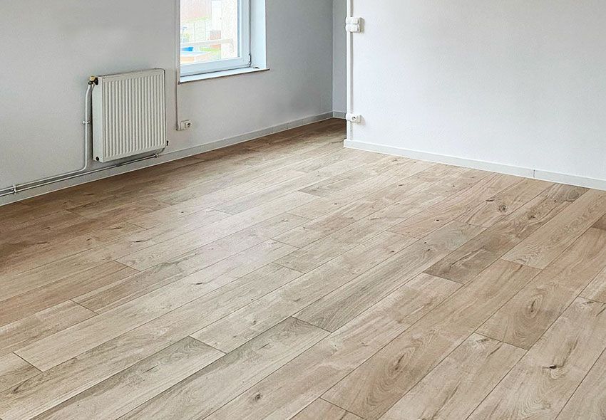 Parquet clair dans une pièce vide aux murs blancs et un radiateur. Une fenêtre est visible en arrière-plan.