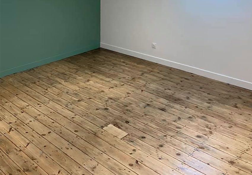 Sol en bois dans une pièce aux murs verts et blancs ; les lames de parquet sont usées et la pièce est vide.