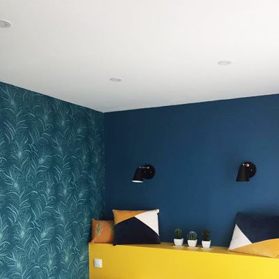 Chambre avec papier peint à motifs turquoise, mur bleu, banc jaune, appliques noires et coussins colorés.