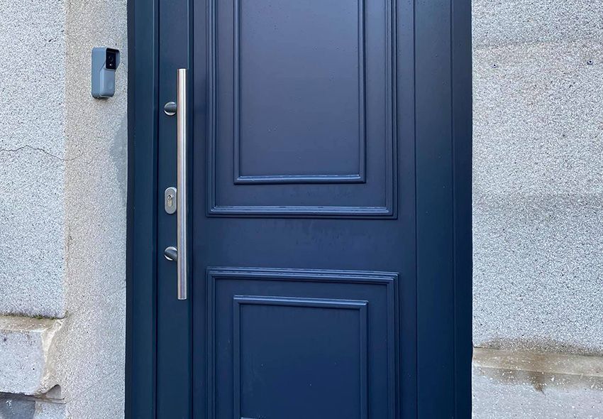 Porte d'entrée bleue avec poignée verticale argentée, encastrée dans un mur en pierre gris clair.