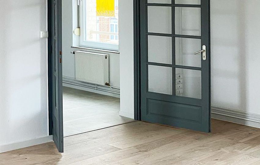 Ouvrez les portes sur une pièce avec parquet clair, murs blancs et une fenêtre avec un radiateur.