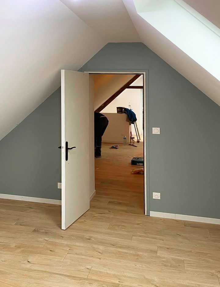 Une porte blanche s'ouvre sur une pièce aux murs gris et au parquet ; une personne travaille à l'arrière-plan.