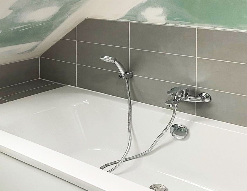 Baignoire avec pommeau de douche et robinet argentés contre un mur carrelé gris.