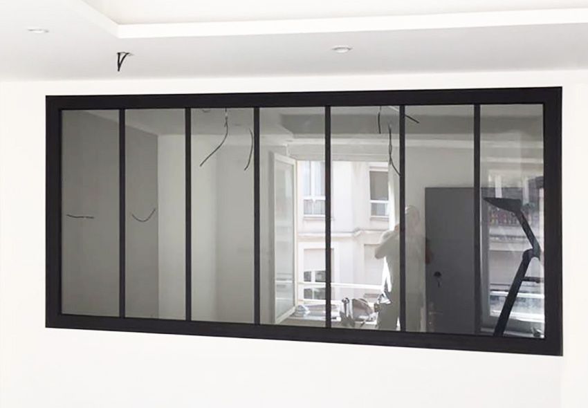 Fenêtre à cadre noir sur un mur blanc. La fenêtre est composée de plusieurs panneaux de verre, reflétant une pièce intérieure avec une fenêtre et une échelle.