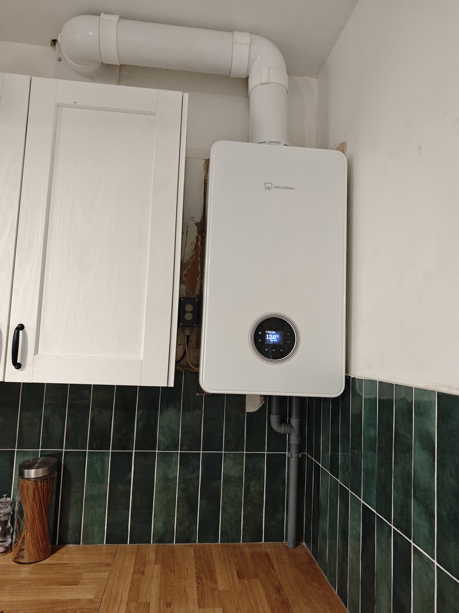 Chaudière à gaz murale blanche avec affichage numérique, tuyau d'évacuation et meuble, dans une cuisine avec crédence carrelée verte.