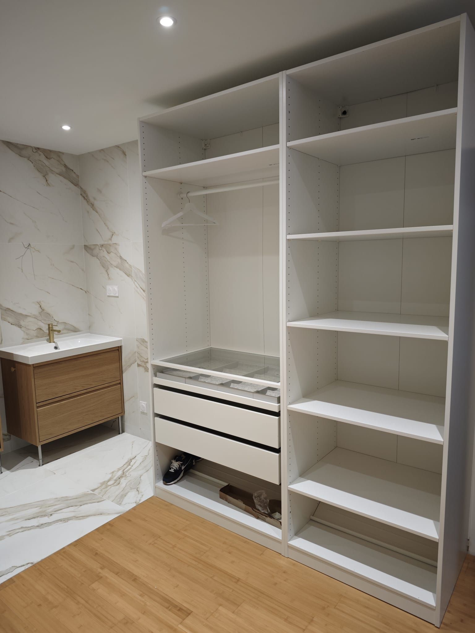 Placard blanc avec étagères et tiroirs à côté d'un meuble-lavabo dans une pièce avec parquet.