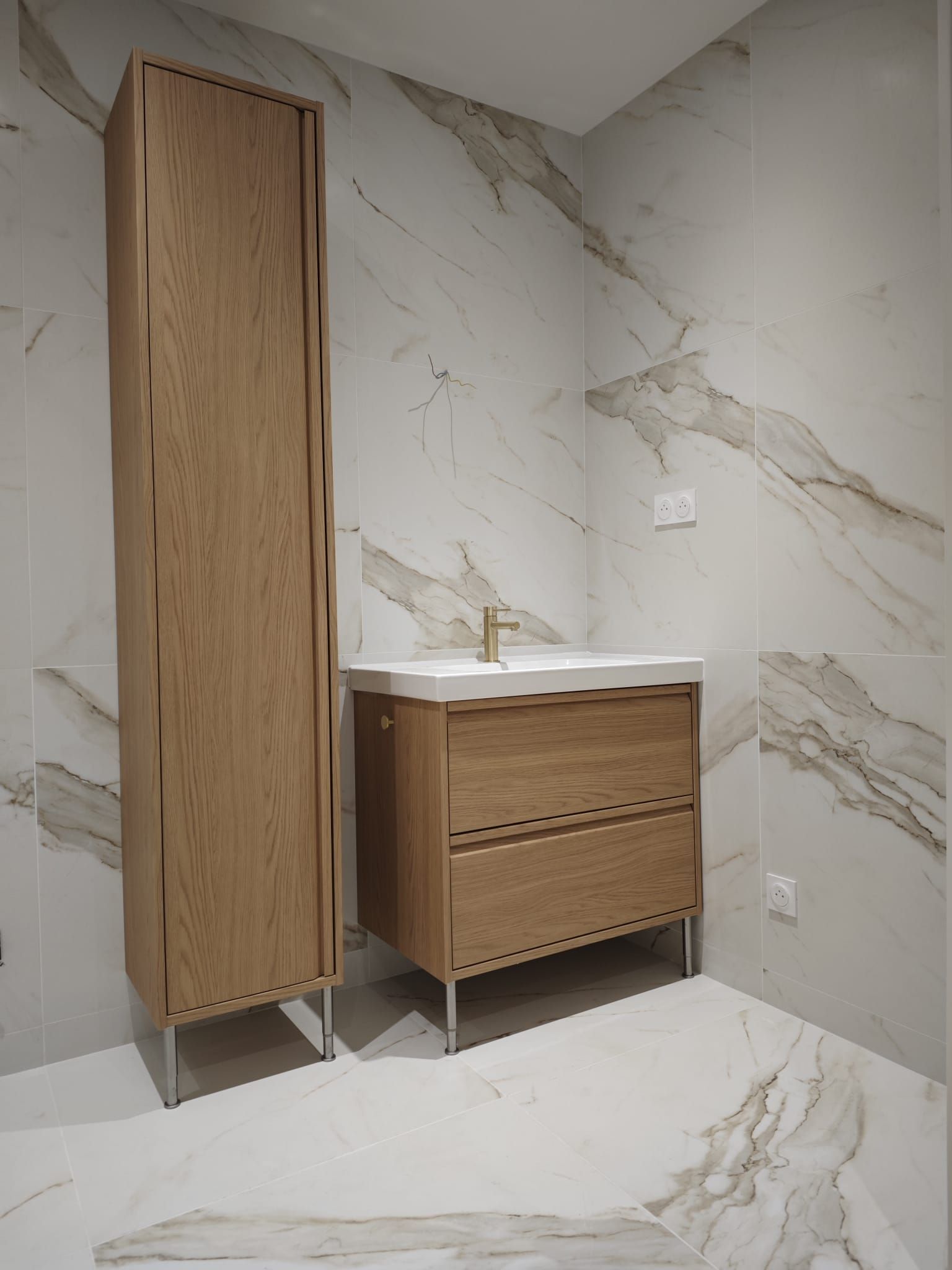 Salle de bains avec meubles en bois clair, lavabo blanc et murs carrelés imitation marbre.