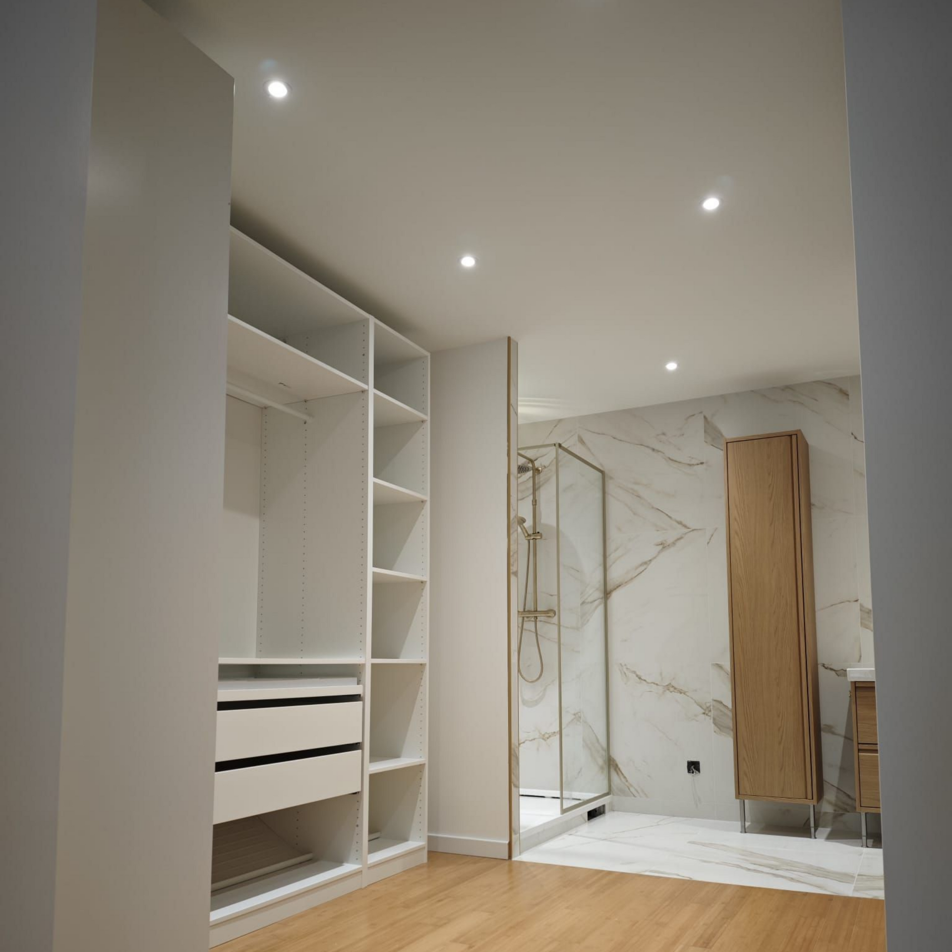 Un placard blanc avec étagères ouvertes et tiroirs, donnant sur une salle de bains aux murs en marbre et à l'armoire en bois.