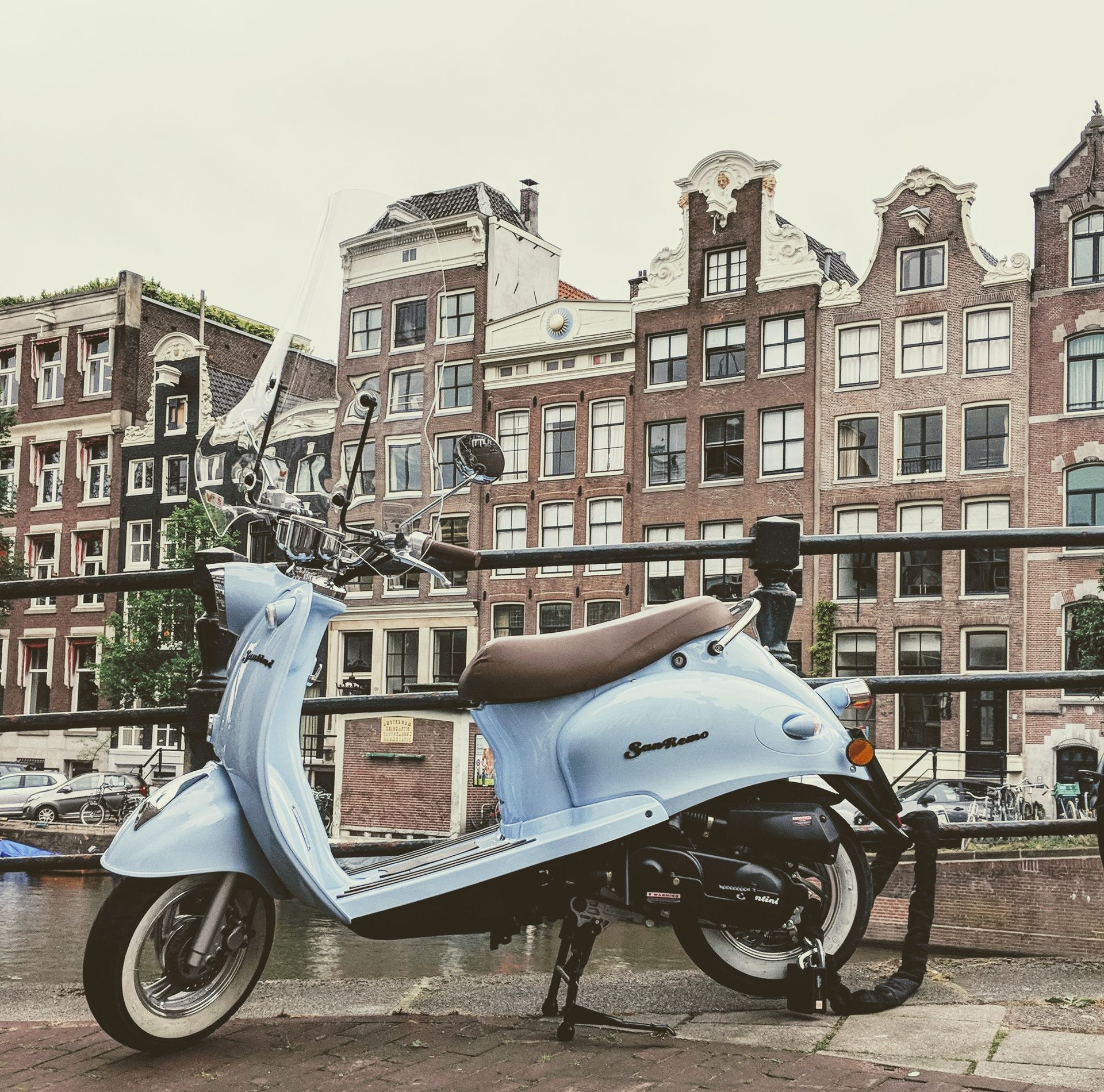 Hellblauer Motorroller, der auf einer Kanalbrücke in Amsterdam geparkt ist, mit historischen Gebäuden im Hintergrund.