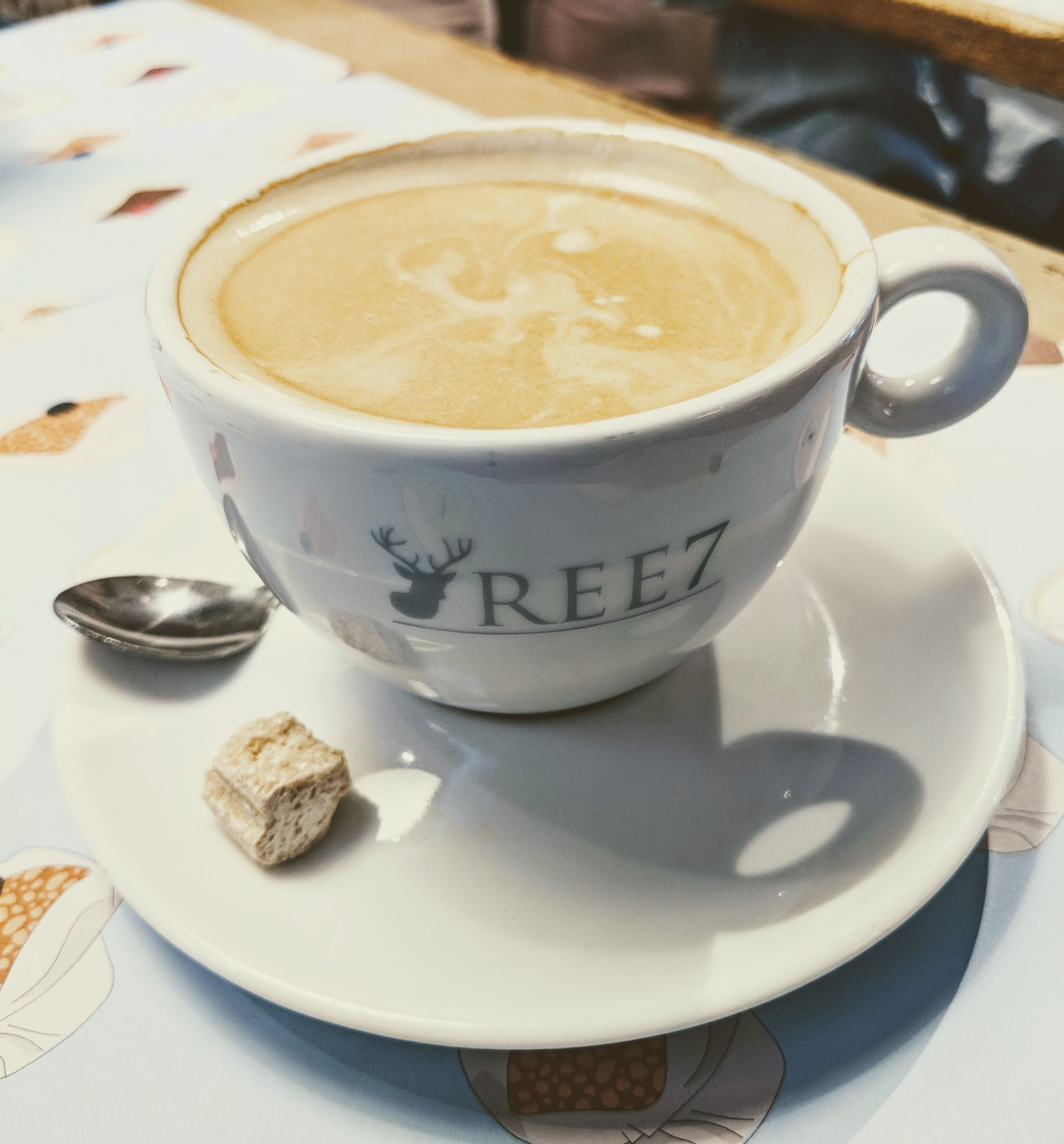 Tasse Kaffee mit einem Zuckerwürfel und einem Löffel auf einer Untertasse. Weiße Tasse mit „REE7“-Logo.